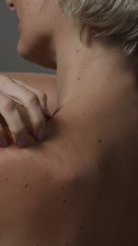 Vertical rear view shot of adult blonde woman scratching bare shoulders suffering from itchy skin on minimal grey background