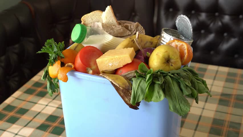 A medium shot captures a blue garbage bin overflowing with a diverse collection of household food waste. The bin contains discarded items such as rotten fruits like apples, bananas, and oranges, along with vegetables like tomatoes and leafy