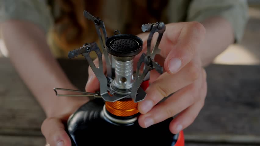 Close-up of a lightweight, compact camping stove on a fuel canister as hands adjust the burner for outdoor cooking, showcasing reliable backpacking gear for minimalist, off-grid adventure, and travel.