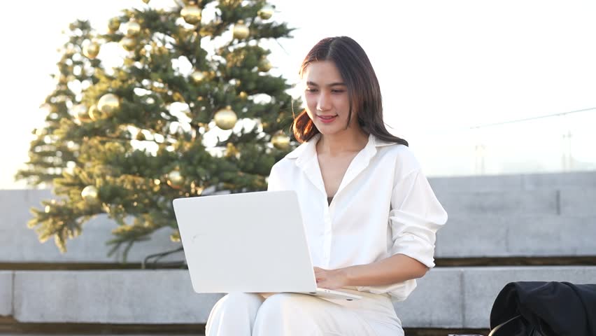 A young woman holding a tablet and jacket checks her phone outdoors, standing near a modern building under bright daylight.
