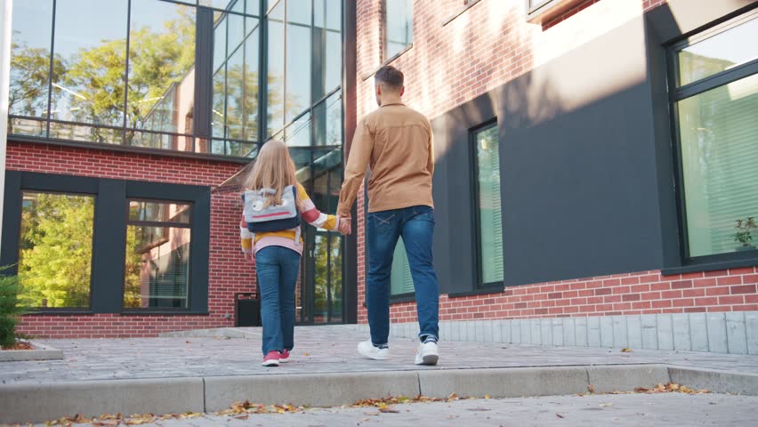 Caucasian father leading daughter with backpack toward school building. Adult male holding child s hand while walking along paved path beside brick walls. Parent accompanying girl on morning drop off.