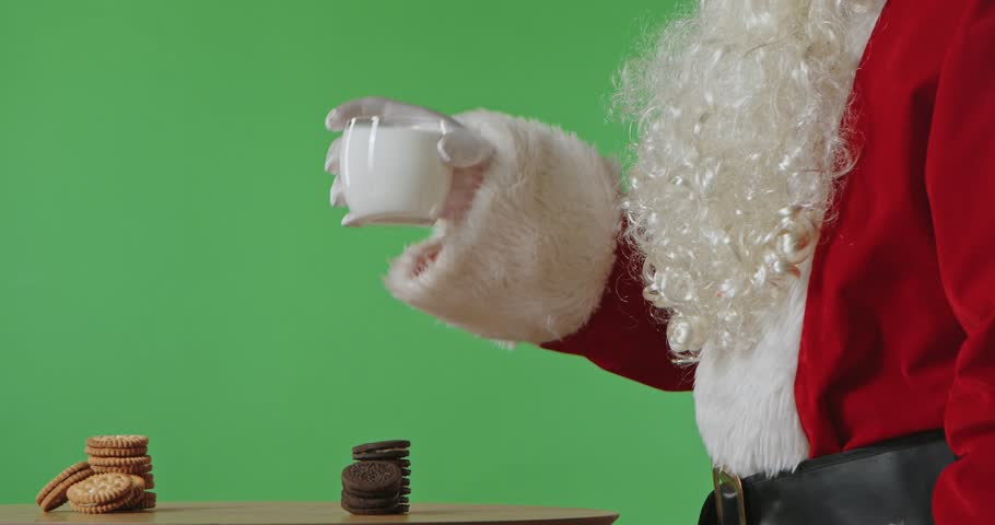Santa Claus is sitting at a table drinking milk with cookies, isolated on a green background.