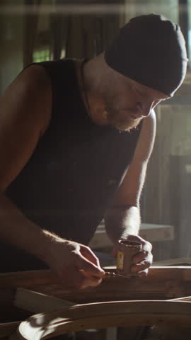 Young man in hat covers flaws with lacquer onto round window frame in carpentry. Focused bearded craftsman restores antique door with material in craft shop