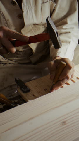 Focused man drives chisel with hammer into solid wood surface. Artisan uses force carefully creating deeper groove with every strike under bright lamp light