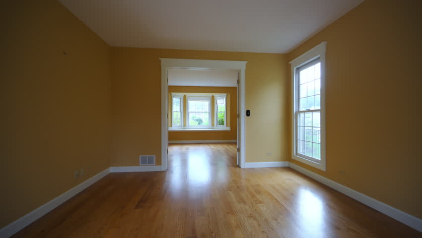 Empty living room in a residential home featuring warm yellow walls, polished hardwood floors, and large windows that fill the space with soft natural light.