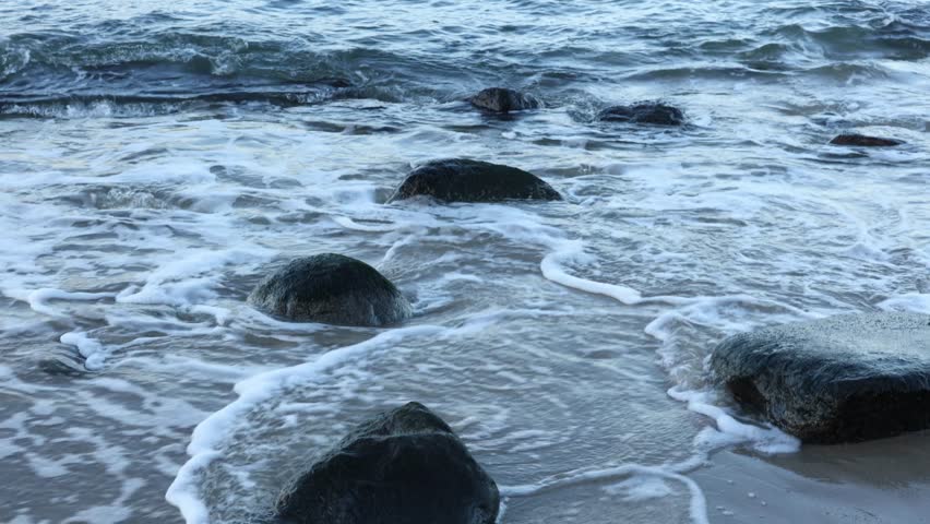 Waves of the sea washing over coastal rocks on sandy beach at sunset. Natural shoreline texture in evening light.