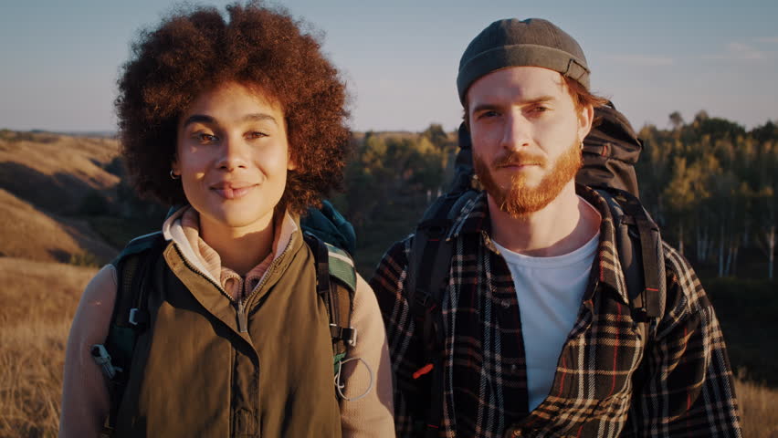 Young mixed race family with Nordic walking poles stands on sunset hill. Happy man and woman tourists hike together at grassy hilly countryside on fall season
