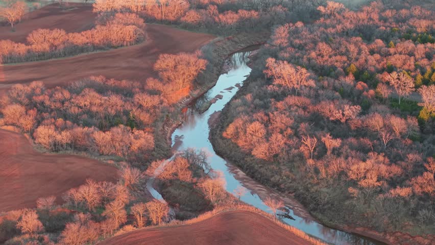 Cinematic golden-hour aerial of a winding river surrounded by glowing winter trees and red earth in Central Oklahoma, captured with smooth dynamic motion.