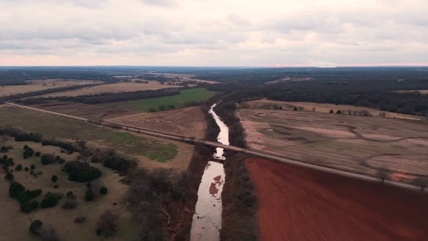 Aerial view of a bridge crossing a winding river between red earth fields and winter farmland in Central Oklahoma, offering a clean documentary-style landscape shot.