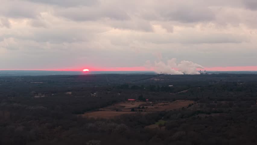 Aerial sunset view of a winter landscape with a distant power plant releasing a steam plume beneath dramatic clouds and a glowing pink horizon in Central Oklahoma.