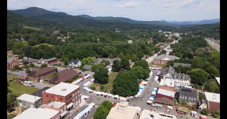 Aerial 4K drone view of downtown Burnsville, North Carolina in summer during a craft fair, showing vendor tents, town streets, and surrounding Blue Ridge Mountain scenery.