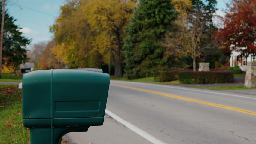 Woman walking to a roadside mailbox, placing letters inside, raising the red flag and leaving along a quiet suburban street on an autumn day. High quality 4k footage