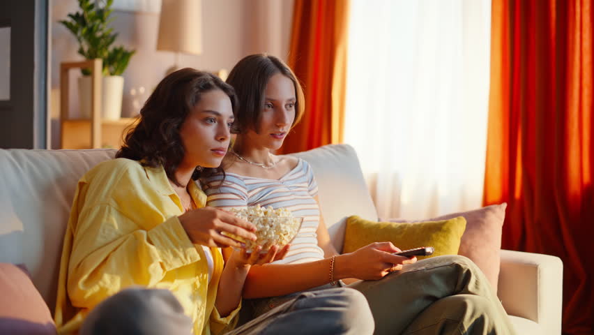 Happy girlfriends watching TV at comfortable home sofa closeup. Relaxed two friends laughing looking comedy show in apartment. Smiling besties eating popcorn enjoying television entertainment together