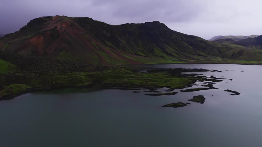 A stunning aerial view of a volcanic crater lake with waterfalls surrounded by rugged terrain and mountains under a cloudy sky in Iceland.