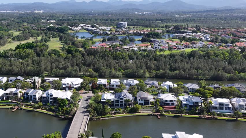 Drone pans across sunny lakeside residential area with modern houses, bridge, greenery, distant hills