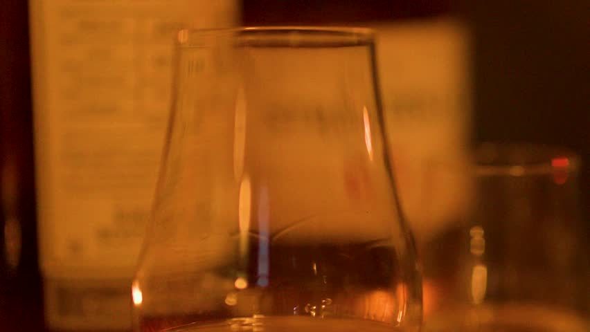 A hand lifts whisky tasting glass from bar, amber liquid, warm evening lighting, shallow focus