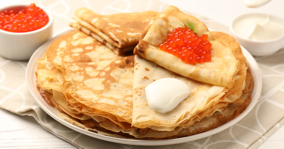Adding sour cream to tasty crepes and red caviar on white table, closeup