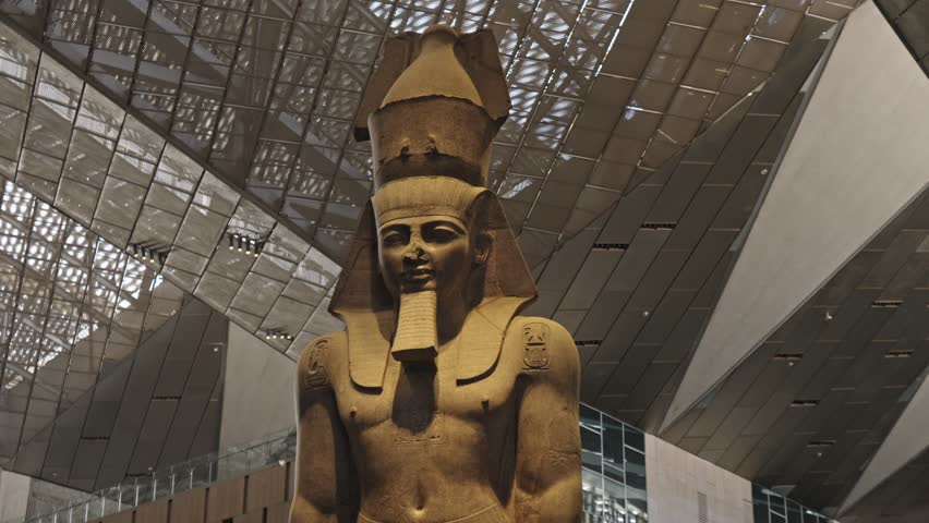 The massive Colossus of Ramesses II standing under the spectacular, contemporary glass and steel roof structure of the Grand Egyptian Museum (GEM) in Giza.