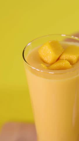 Close-up of hand topping mango smoothie with fruit cubes on bright yellow background