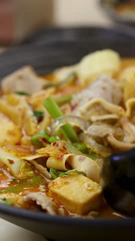 Close-up of hand stirring steaming spicy hotpot soup with tofu, meat, vegetables, shallow focus