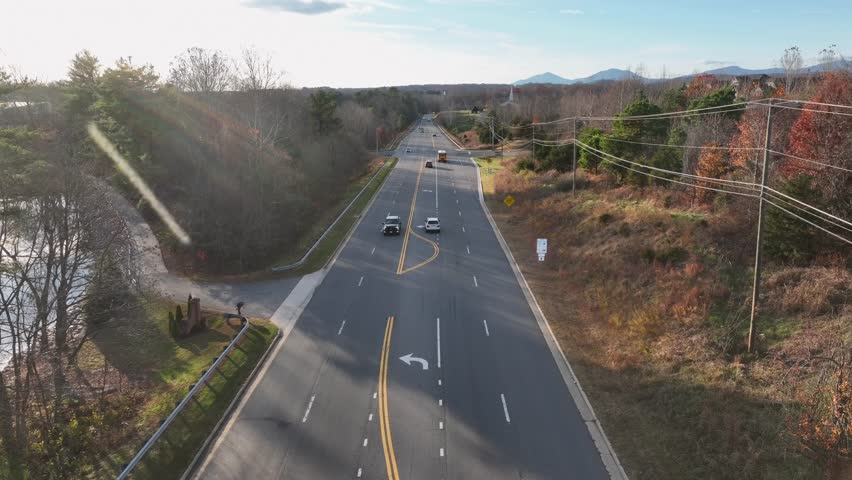 America forest roadway in autumn with warm sunlight, rolling hills, yellow school bus and scattered homes along quiet rural corridor surrounded by colorful trees. Aerial wide shot.