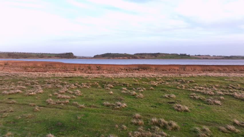 A low-flying drone captures an autumn meadow with tall grass beside calm water during a warm evening sunset in Denmark’s Jutland region.