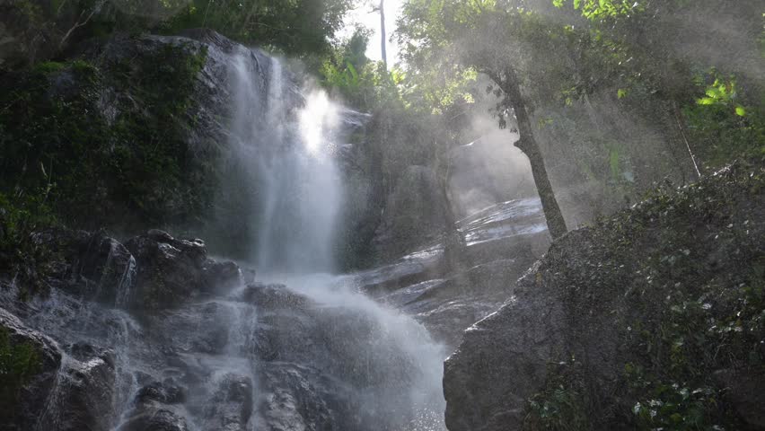 Sunbeams shining through the mist of a beautiful waterfall flowing over rocks in a green forest