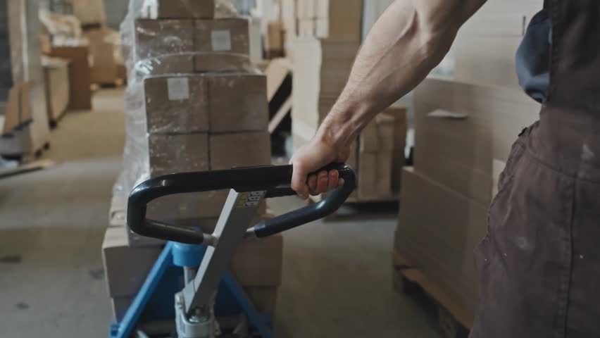 Cropped shot of a male worker pulling a cart loaded with cardboard boxes, highlighting warehouse work, logistics, and manual labor in an industrial setting.
