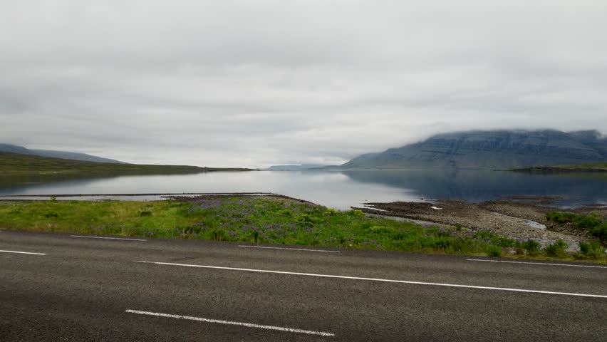 Aerial drone footage of a tranquil paved road beside a serene fjord with misty mountain backdrop near Seyðisfjörður, East Iceland. Captures the calm landscape and cloudy sky.