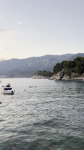 Scenic harbor bay at golden hour sunset. Sun rays break through clouds over chain of islands casting glow on calm water reflecting sky. Boats anchored near shore, visible mooring lines. Houses at cape