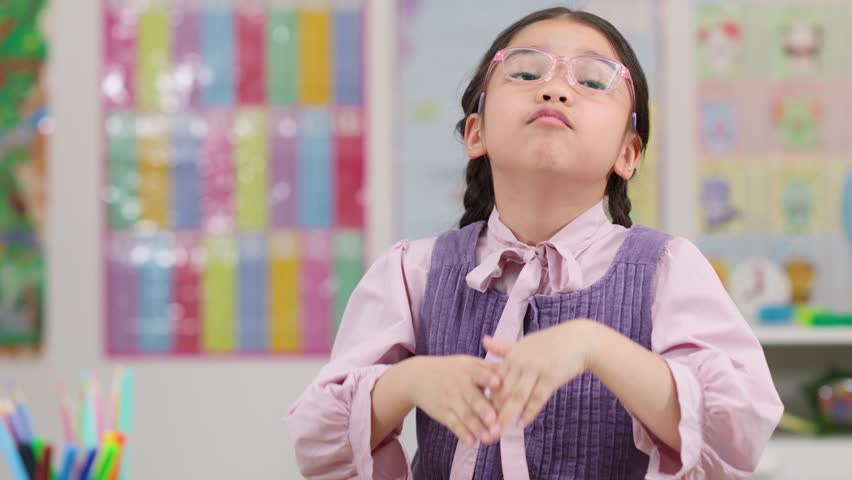 Young girl with braids and glasses pleads at desk in colorful, softly lit classroom.