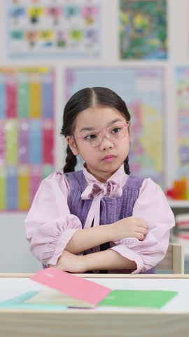 Young girl with braids and glasses sits at desk, pondering thoughtfully in bright classroom setting
