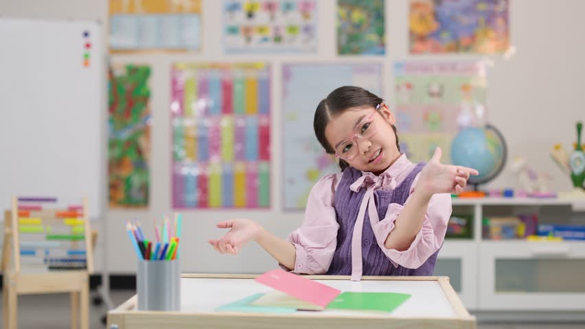 Young girl with glasses smiles and laughs at desk in brightly lit classroom, static camera