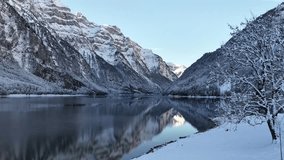 Cold, serene winter panorama of Klöntalersee, Glarus, Switzerland. A snow-dusted tree frames the lake's still, dark surface, which reflects the massive, steep alpine peaks. Quiet wilderness escape. - Powered by Shutterstock - Get 15% off with code: PIKWIZARD15