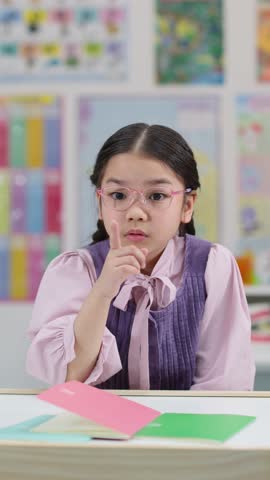 Young girl signals for quiet in classroom, wearing glasses and braids, bright indoor lighting