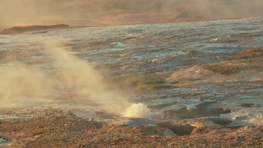 A close up shot of a bubbling mud pot and a steam vent in the Hverir geothermal area of Iceland. The rugged, colorful landscape is illuminated by the warm light of the setting sun, highlighting the in