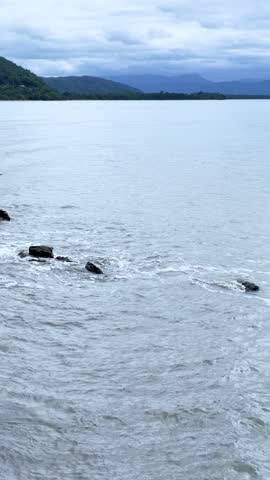 Camera pans over rocky shoreline as ocean waves wash against rocks, lush green hills background