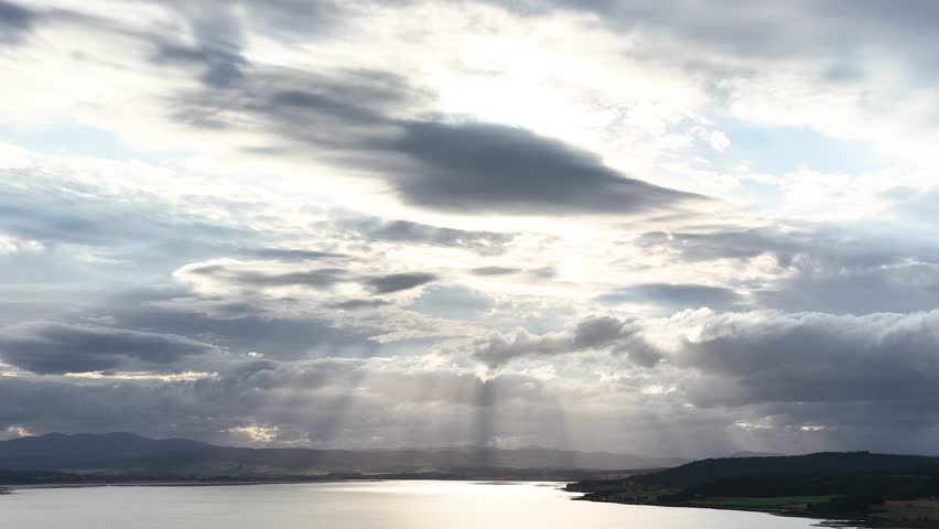Dramatic sunbeams pierce clouds above tranquil water and hills, wide landscape, soft evening light