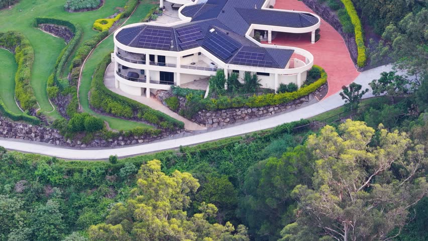 Drone circles hillside luxury house with solar panels, lush landscaping, and curved driveway, daylight