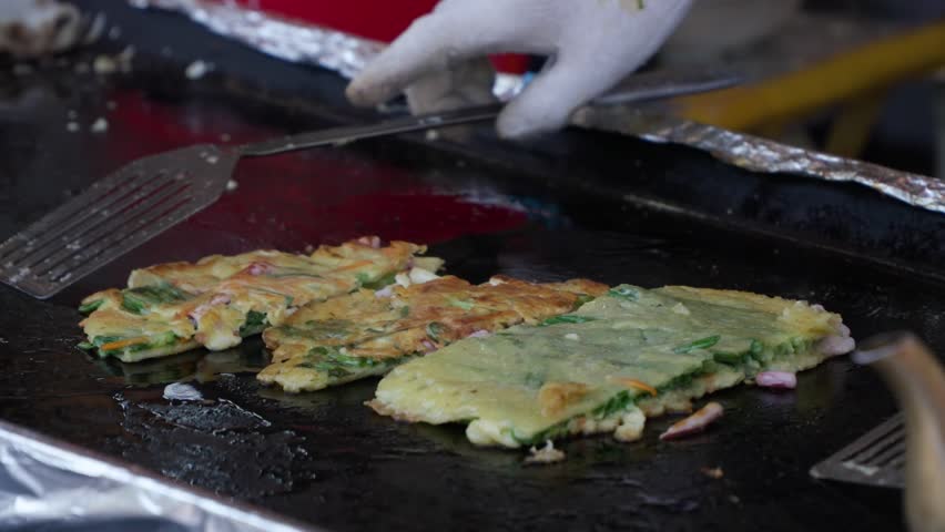 Close-up of a vendor's gloved hands using spatulas to flip and cook savory Korean street food pancakes on a large, shimmering metal grill at a local outdoor market