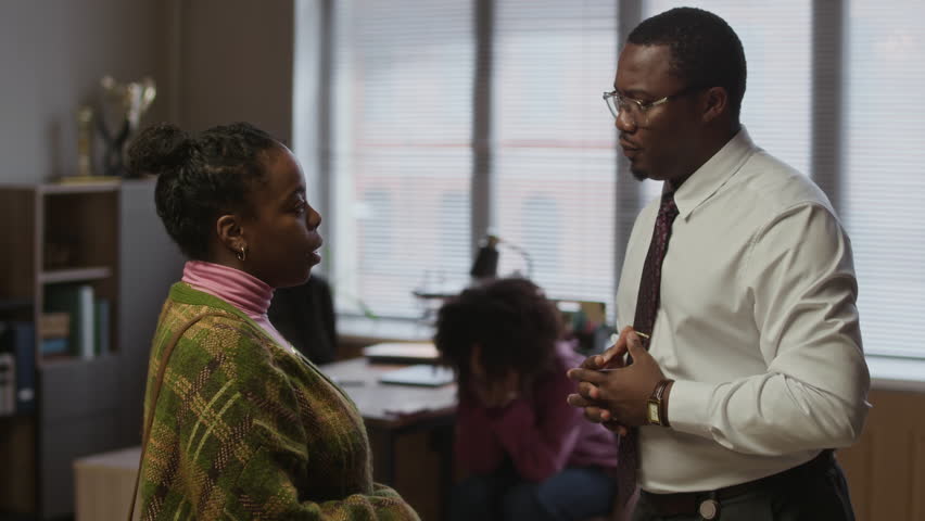 Medium shot of young African American male principal talking to mother of female teenager with misbehavior in school office