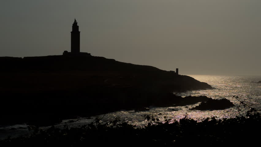 Silhouetted lighthouse on a rocky coast at dusk, with a sunlit sea and space reserved for text.