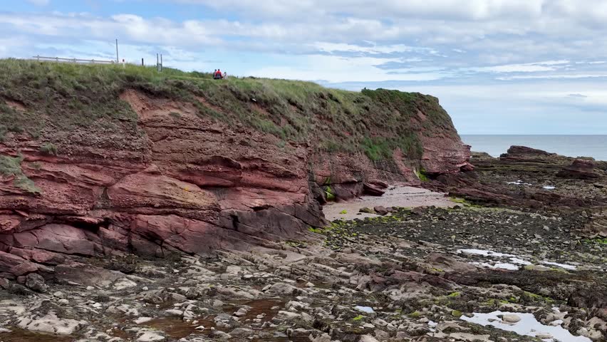 Camera slowly pans revealing red sandstone cliffs, rocky shore, seaweed, and overcast coastal landscape