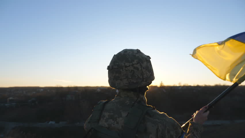 Military man in uniform waving flag of Ukraine against background of sunset. Male ukrainian army soldier lifting national banner at hill in honor of the victory against russian aggression. Rear view