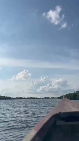 POV shot from a small wooden boat moving through a wide river or lake under a bright blue sky with white cumulus clouds. Scenic view of tropical nature and water transportation.