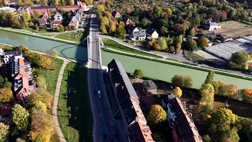 German town scene with a canal bridge, residential houses, autumn trees and long building covered in rooftop solar panels, illuminated by midday sunlight. Aerial top down shot.