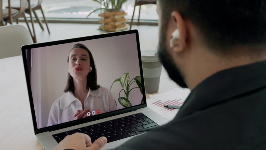Indian businessman with headphones listens to woman financial counselor on video call in office. Young entrepreneur discusses strategy of company growth remotely via laptop indoors