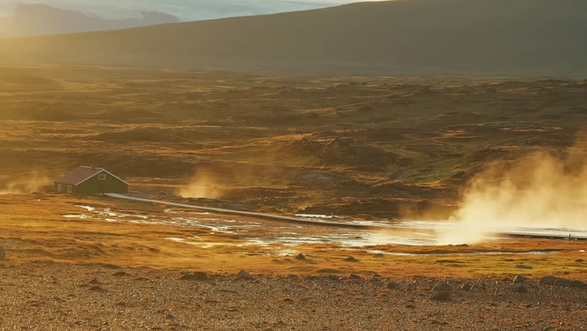 A stunning wide shot of a geothermal landscape in Iceland during a beautiful sunset. A geyser erupts with a powerful burst of steam and hot water, while a small, traditional wooden house sits in the d