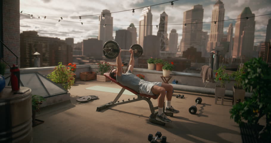 Athletic Man Lies on an Incline Bench, Lifting a Loaded Barbell Upward With Focused Effort, Performing Slow, Controlled Bench Press Repetitions During a Solo Strength Workout on Rooftop