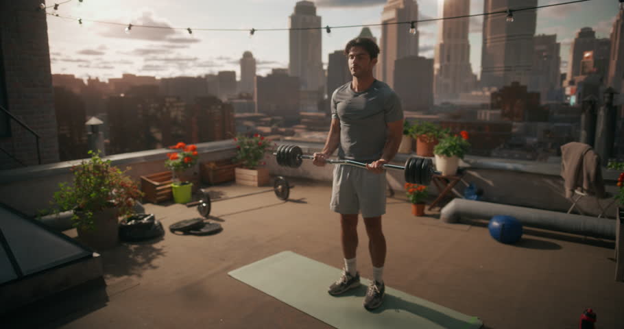 Young Man Stands Doing Slow Ez Bar Curls, Training Arm Strength and Fitness With Light Gym Gear Around Him, Focusing on Exercise and Everyday Health Care. Rooftop City View